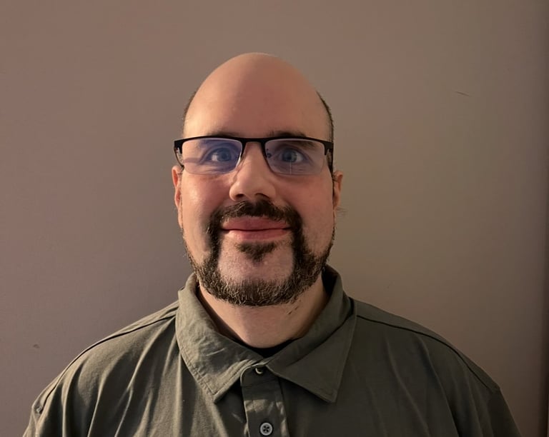 Portrait of a bald man with glasses and a dark beard wearing an olive-colored collared shirt against a neutral background